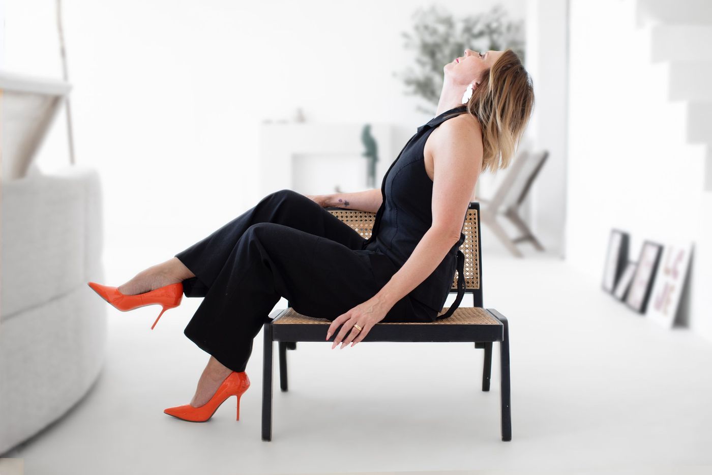 Woman in a black jumpsuit and orange heels leaning back on a cane bench in a bright white room – personality branding by wo0