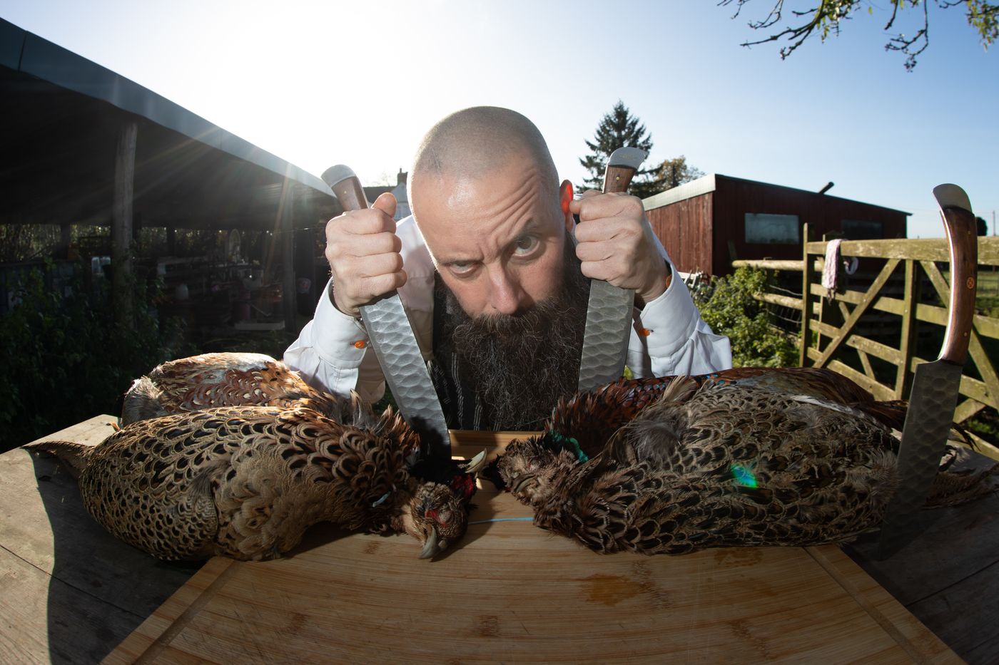 Bearded butcher holding two knives over a pair of pheasants outdoors – brand photography for men by wo0