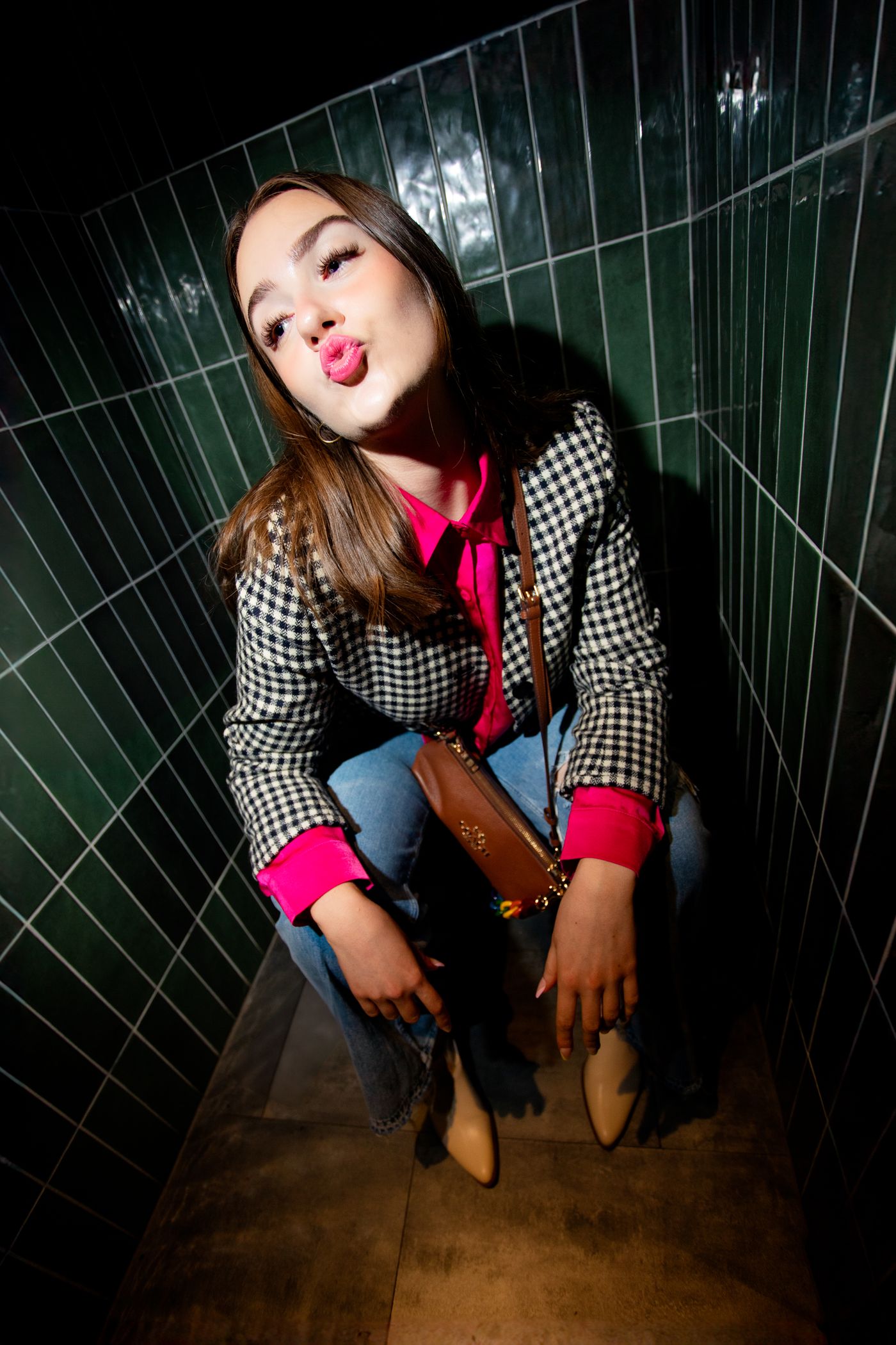 Woman in a houndstooth jacket and pink shirt crouching against green tiles, blowing a kiss – personality branding by wo0
