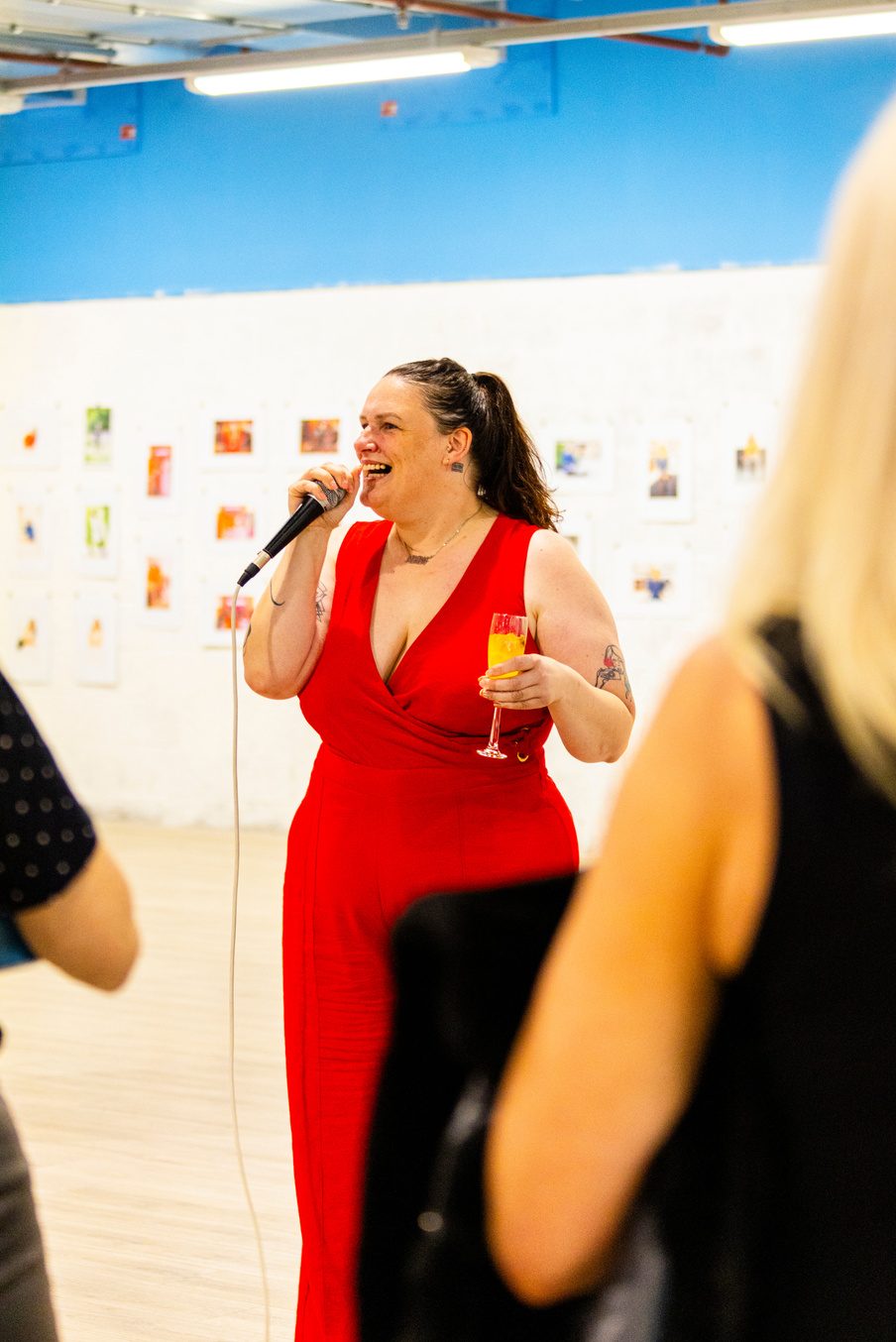Wendy ‘wo0’ Gannon in a red jumpsuit laughing into a microphone with a drink in her hand at a photography exhibition