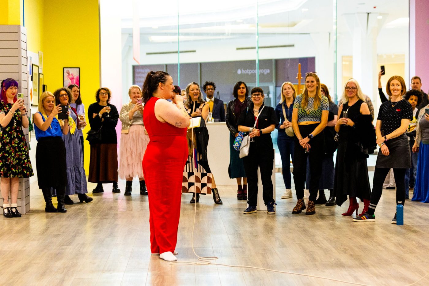 Wendy ‘wo0’ Gannon in a red jumpsuit speaking into a microphone to a laughing crowd at an exhibition launch